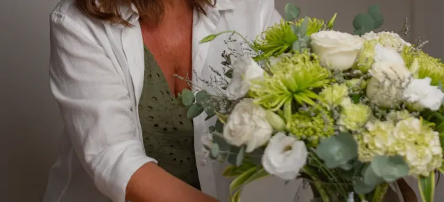 Florista de camisa branca segurando um arranjo de rosas brancas, lisiantos brancos e crisântemos verdes com eucalipto
